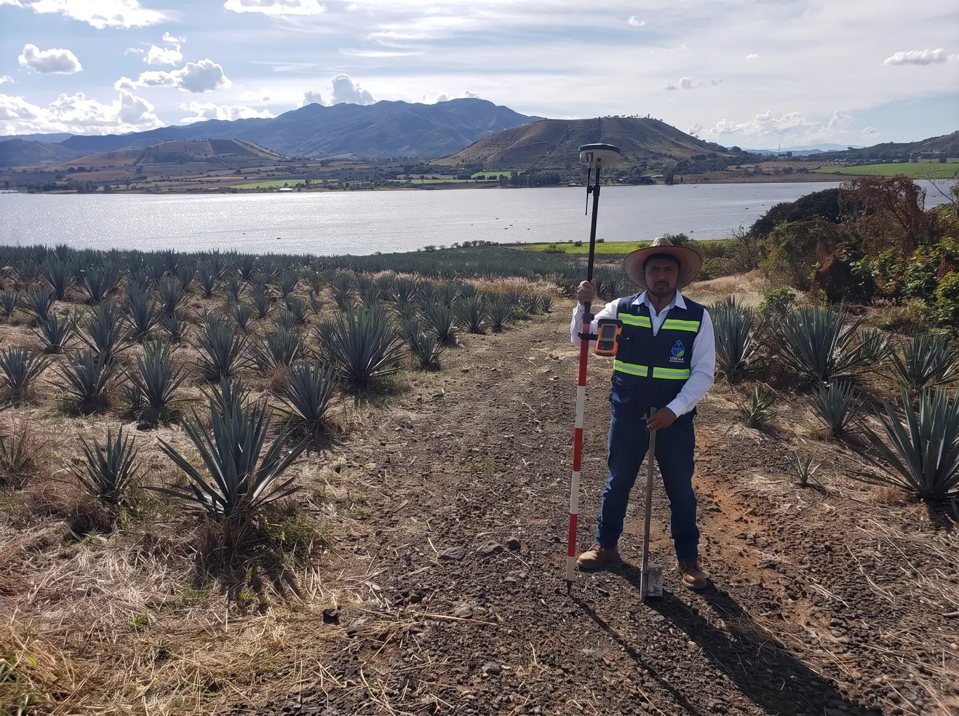 Brigadista con chaleco y bastón GNSS en agave junto al lago
